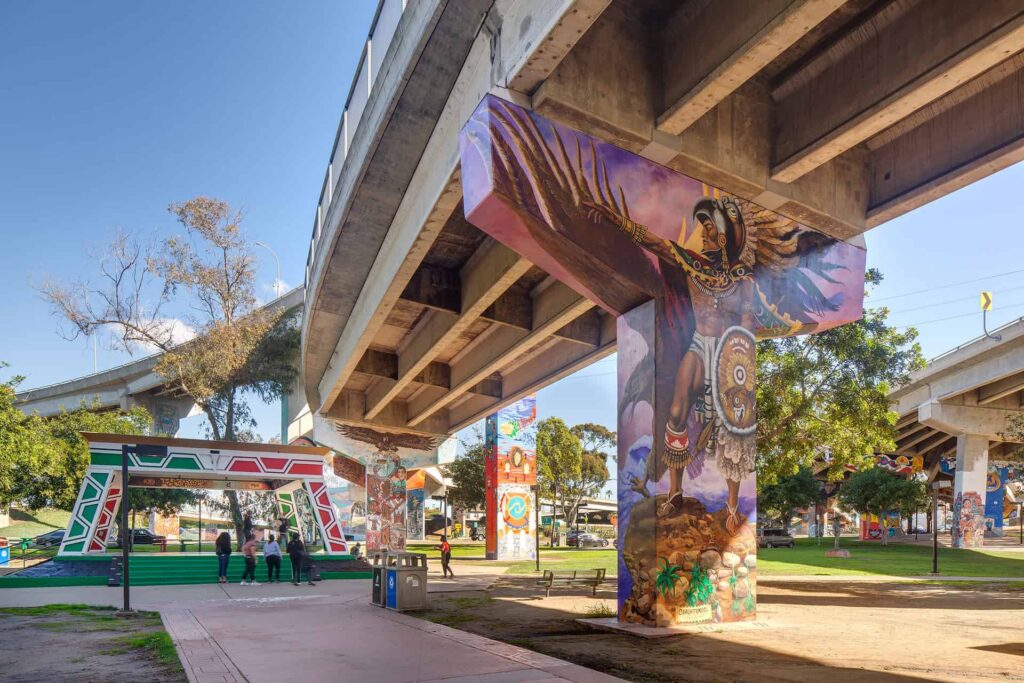 A freeway bridge runs atop a public park. Colorful murals are painted on the concrete pillars that hold up the bridge.