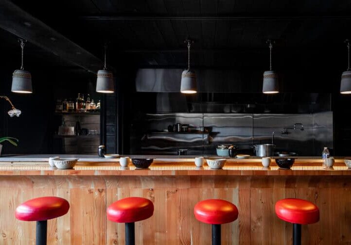 The inside of a ramen restaurant that includes black walls, a wooden bar, and bright red bar stools.