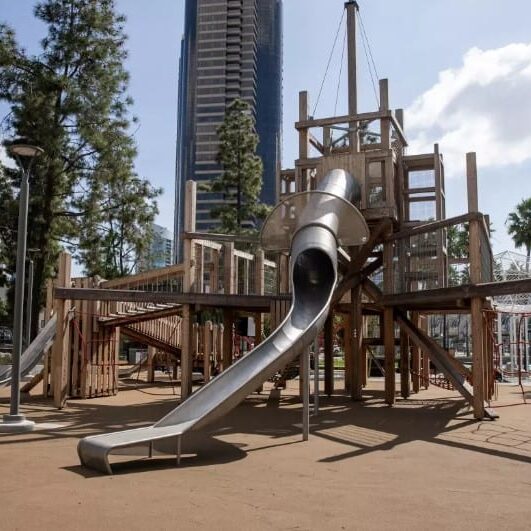 A large, modern play structure is set against the backdrop of downtown high rise buildings.