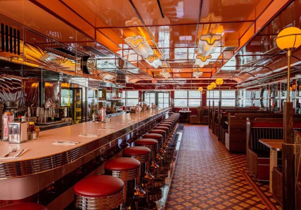 Inside of an upscale diner with warm lighting, tiled floors, a long countertop, and classic red counter stools.