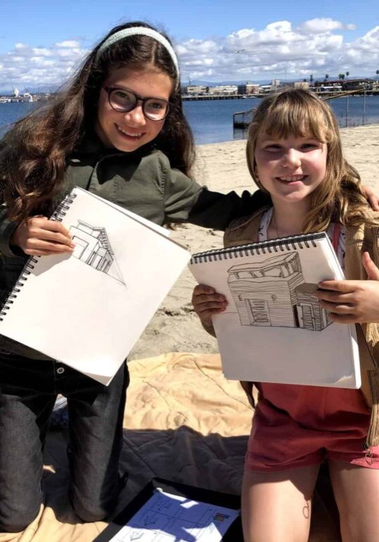 Two young girls kneel on the beach, holding up their sketches